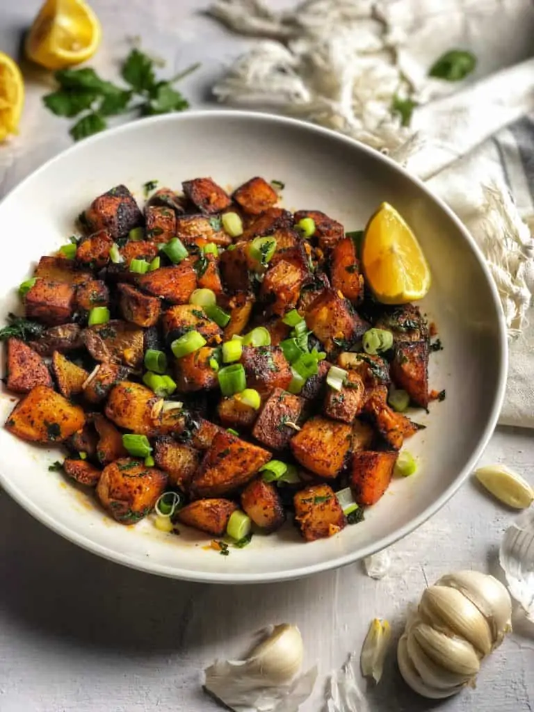 A bowl of seasoned roasted potatoes, inspired by batata harra, garnished with chopped green onions. Lemon wedges complete the vibrant scene.