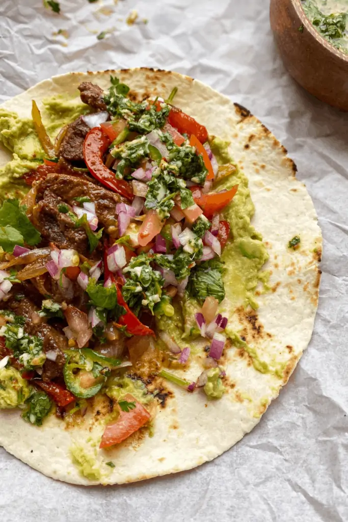 steak fajita with diced red onion, parsley, guacamole, steak, and red bell peppers on a tortilla on a parchment paper