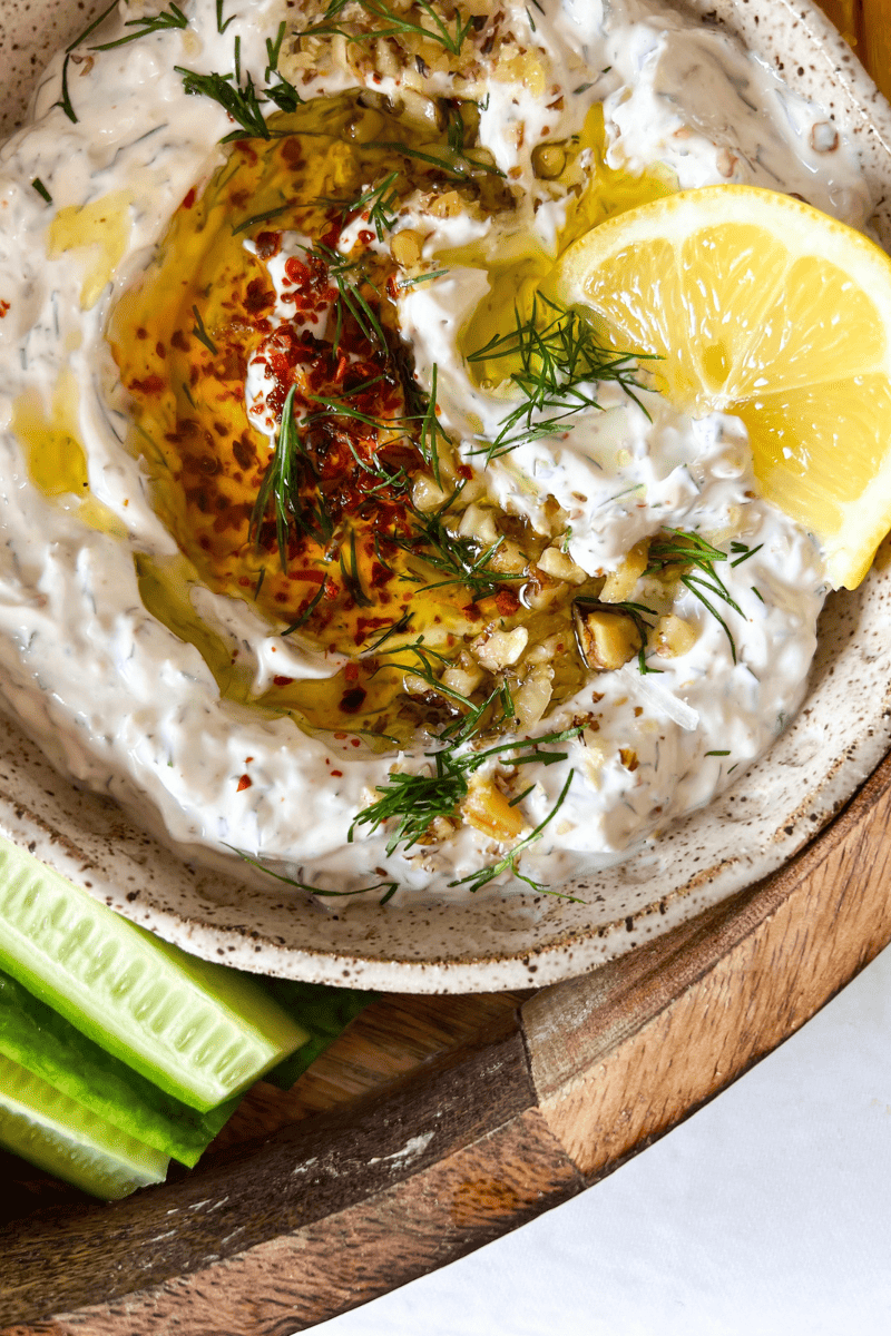 labneh dip with a slice of lemon on top, walnuts, dill, aleppo pepper and drizzle of olive oil served with cucumbers