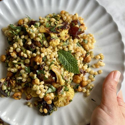 A hand holds a white plate filled with Roasted Cauliflower and Couscous Salad, featuring chickpeas, herbs, dates, and roasted vegetables, all beautifully garnished with a fresh mint leaf.