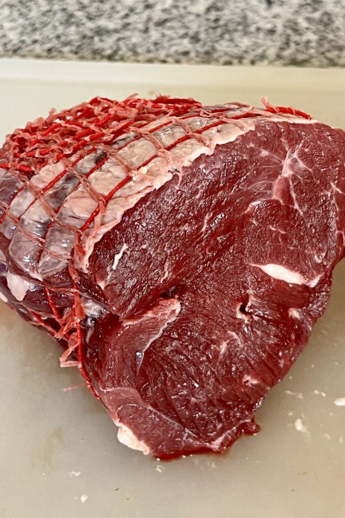 A large cut of raw lamb with a red netting partially removed, for middle eastern lamb skewers (lamb shish kebab). It's resting on a cutting board with a granite countertop in the background