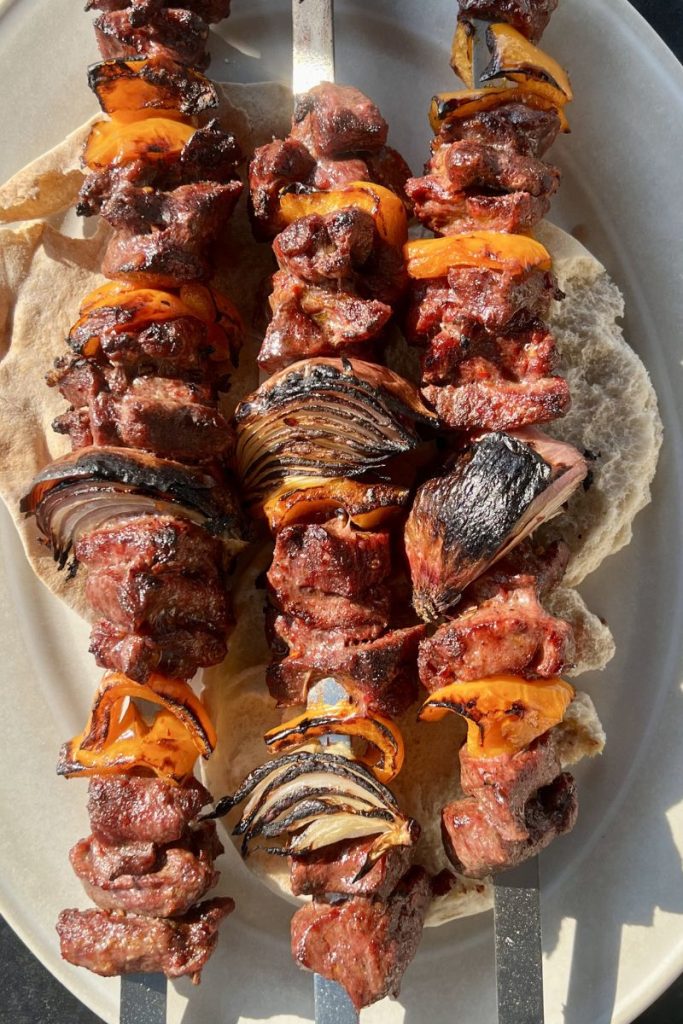 Three skewers with grilled beef and lamb shish kebab, orange bell peppers, and charred onions on thin pita bread, served on a round beige plate.