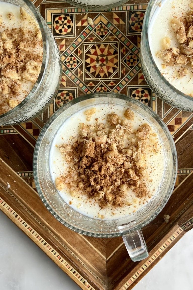 Three glass cups of mahalabia (muhellabi) milk pudding topped with chopped walnuts and ground cinnamon are placed on a decorative wooden tray with geometric patterns.