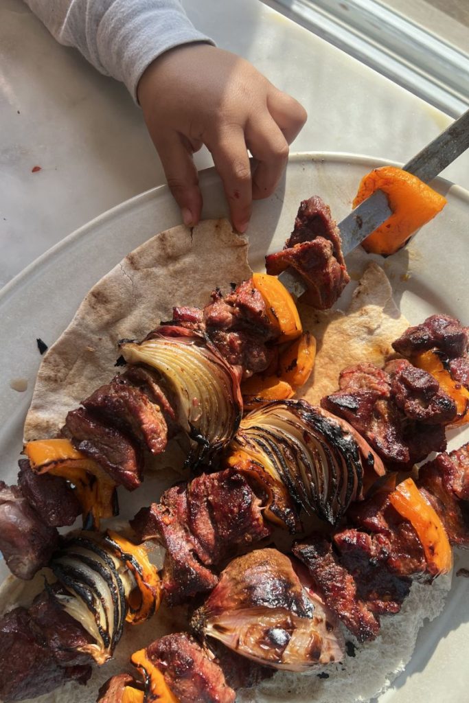 A child’s hand reaches for flavorful Middle Eastern lamb skewers also called lamb shish kebab and vegetables on thin pita bread, with sunlight illuminating the food on a white plate.
