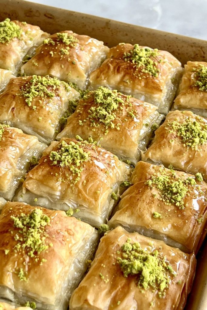 Rows of homemade golden-brown pistachio baklava pieces, each topped with chopped pistachios, arranged neatly in a baking tray.