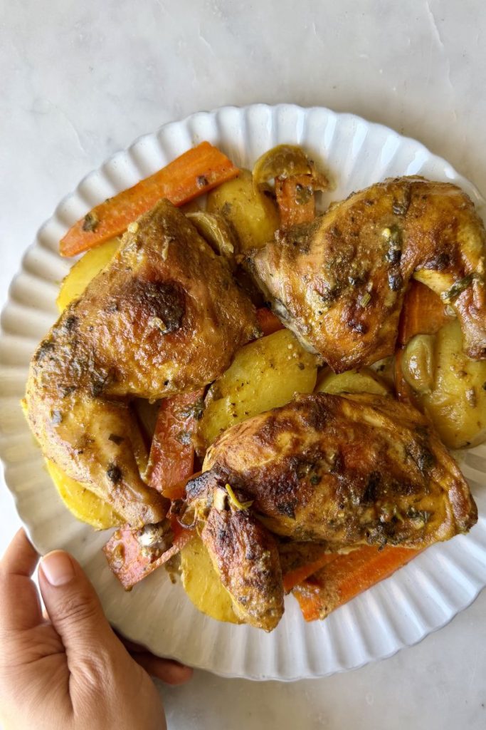 hand holding a white plate of Moroccan roasted chicken with potatoes and carrots